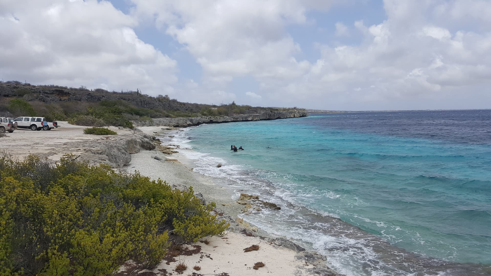 Duikstek aan de rotsachtige westkust van Bonaire met turquoise water, duikers in de branding, droge struiken en geparkeerde auto's