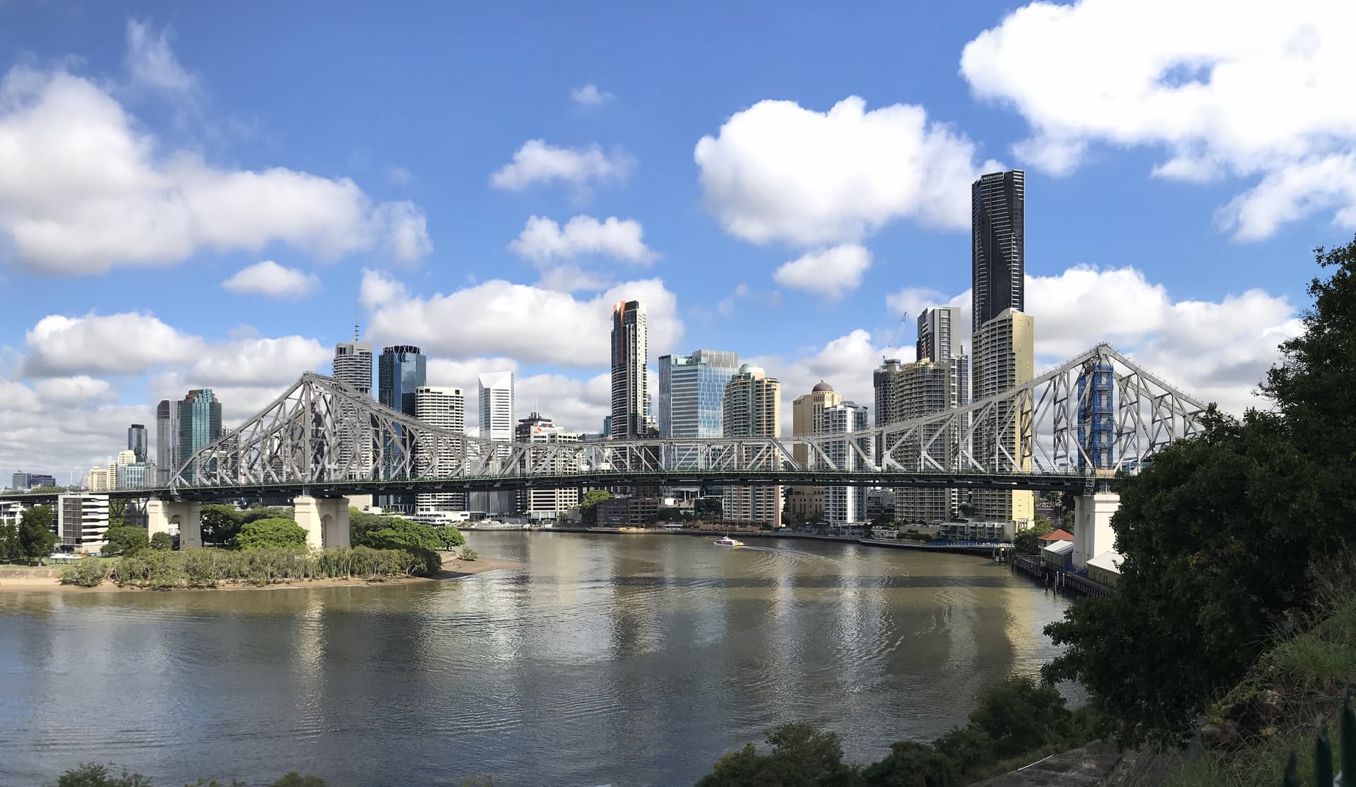 De stalen Story Bridge over de bruine Brisbane River met daarachter de wolkenkrabbers van het centrum van Brisbane onder een zonnige lucht met stapelwolken