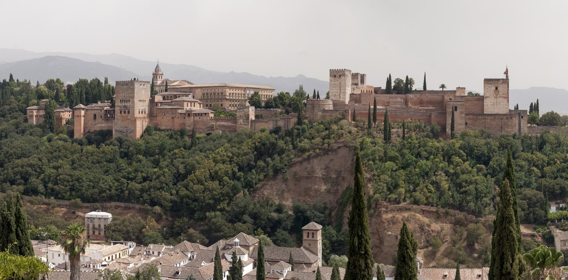 De rode muren en torens van de Alhambra op een beboste heuvel boven Granada, met bergen op de achtergrond