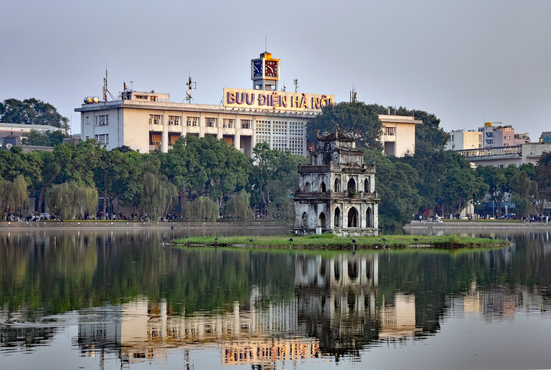 De Schildpaddentoren (Thap Rua) op een eilandje in het Hoan Kiem-meer in Hanoi, weerspiegeld in het stille water, met het koloniale postkantoor erachter
