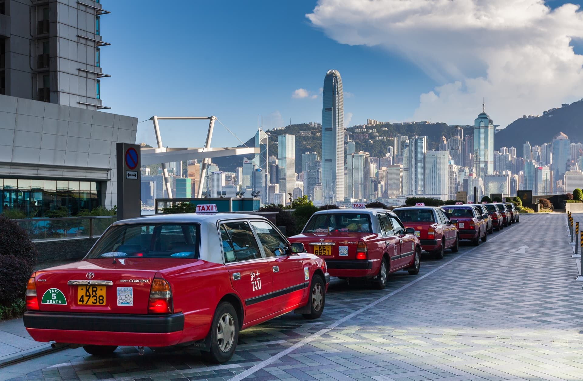 Rij rode taxi's aan de waterkant van Kowloon, met aan de overkant van Victoria Harbour de wolkenkrabbers van Hong Kong Island en de heuvels erachter