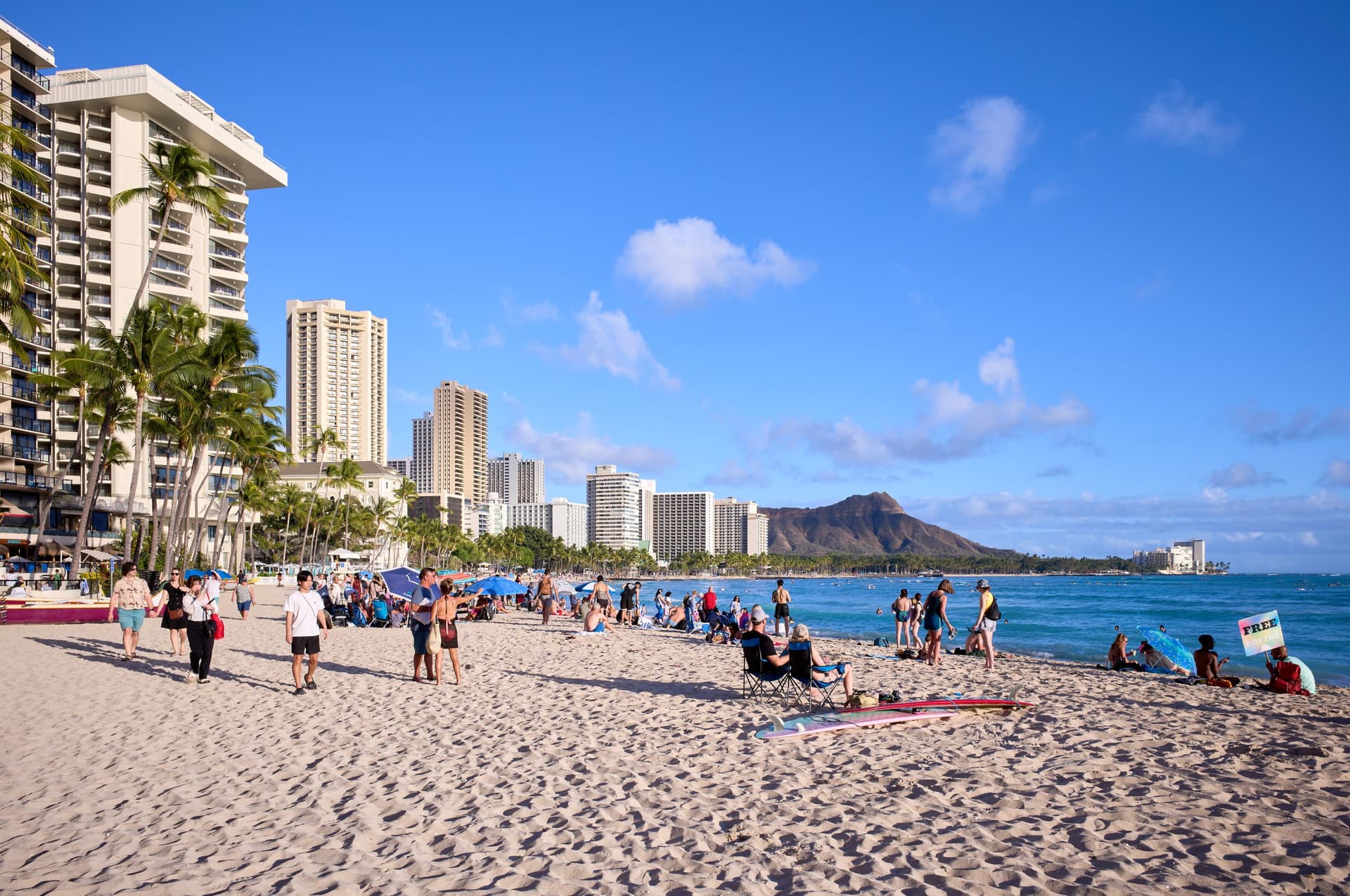 Het zandstrand van Waikiki vol badgasten, met hoteltorens en palmen links en de krater Diamond Head aan de horizon onder een blauwe lucht