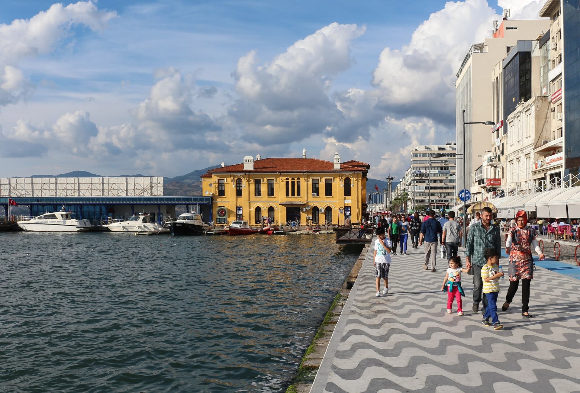 Wandelaars op de kade van de Kordon in İzmir met een geel historisch gebouw aan het water