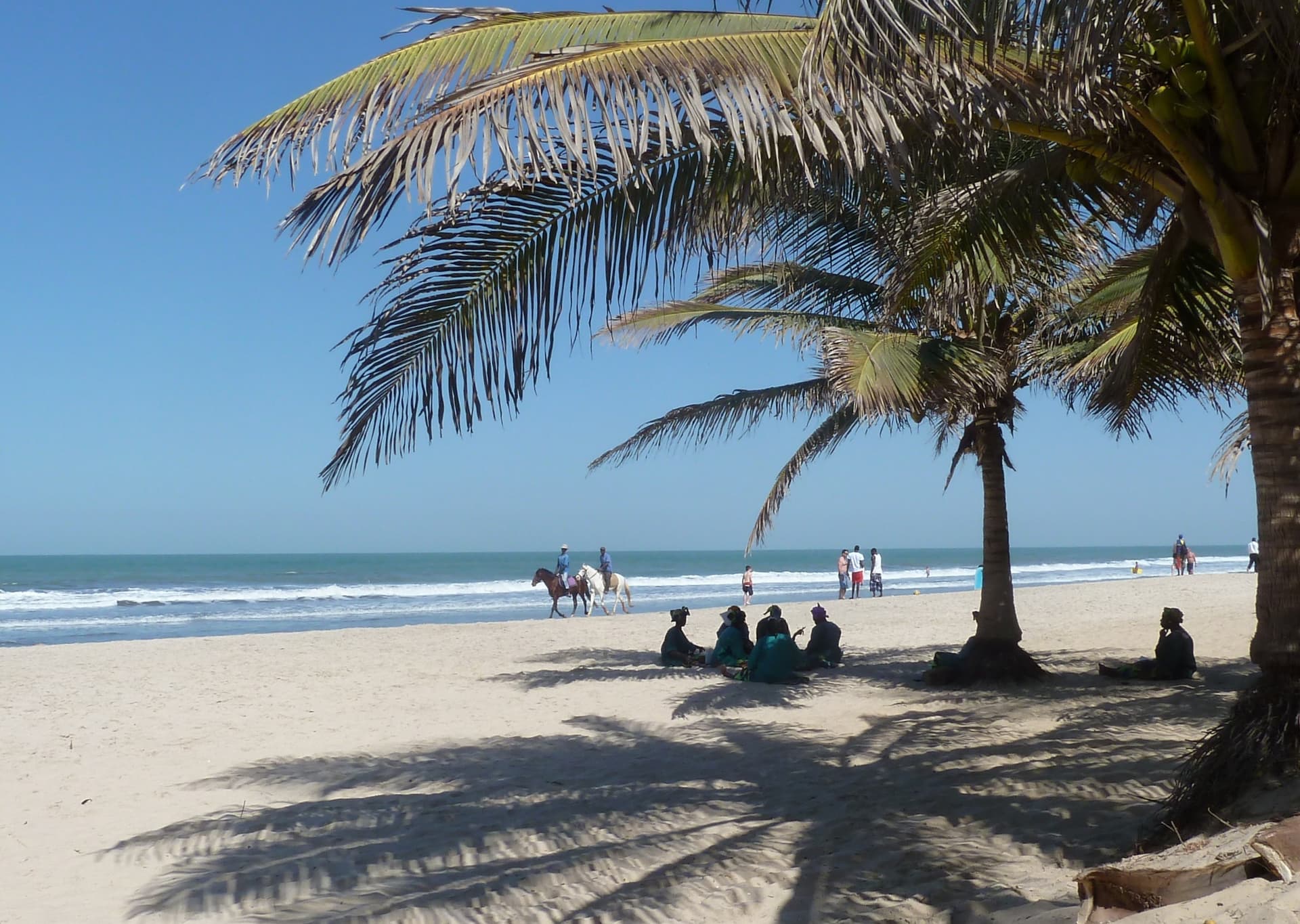Wit zandstrand bij Kotu met palmbomen, ruiters langs de branding en mensen in de schaduw onder een blauwe lucht