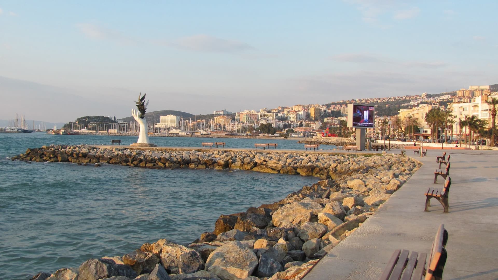 De kustpromenade van Kuşadası met een golfbreker en een wit vredesbeeld in zee, met de stad langs de baai op de achtergrond