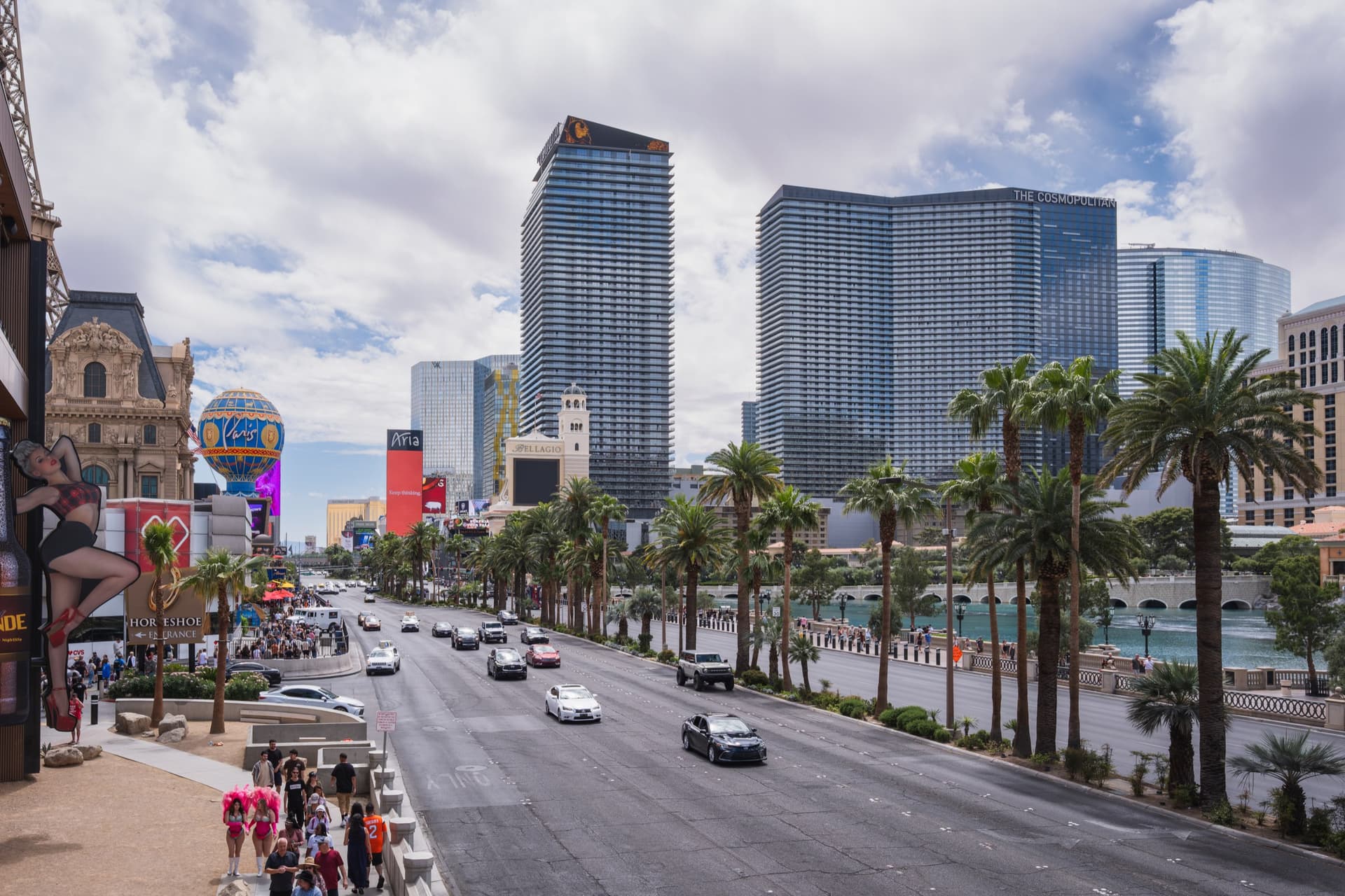 Las Vegas Boulevard overdag met palmbomen, de Bellagio-fontein en de torens van The Cosmopolitan onder een lucht met stapelwolken