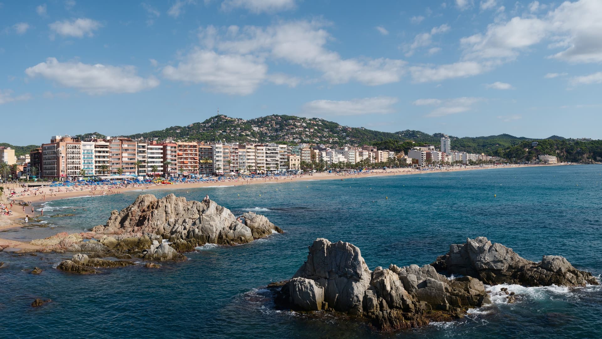 Het lange stadsstrand van Lloret de Mar met hotels en groene heuvels, gezien over rotsen in de zee