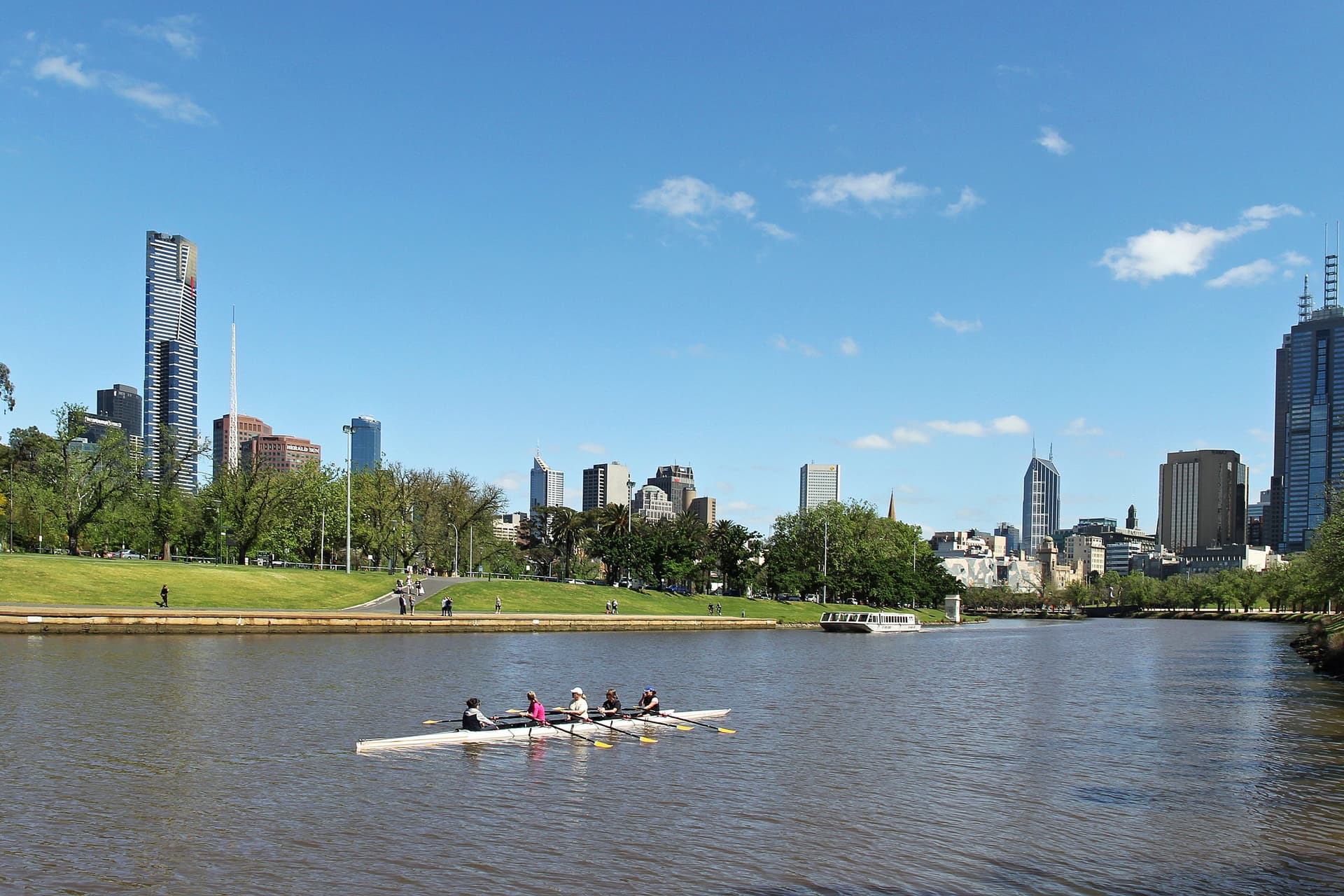 Roeiers op de Yarra in Melbourne, met groene oevers, parken en de torens van het centrum op de achtergrond onder een blauwe lucht