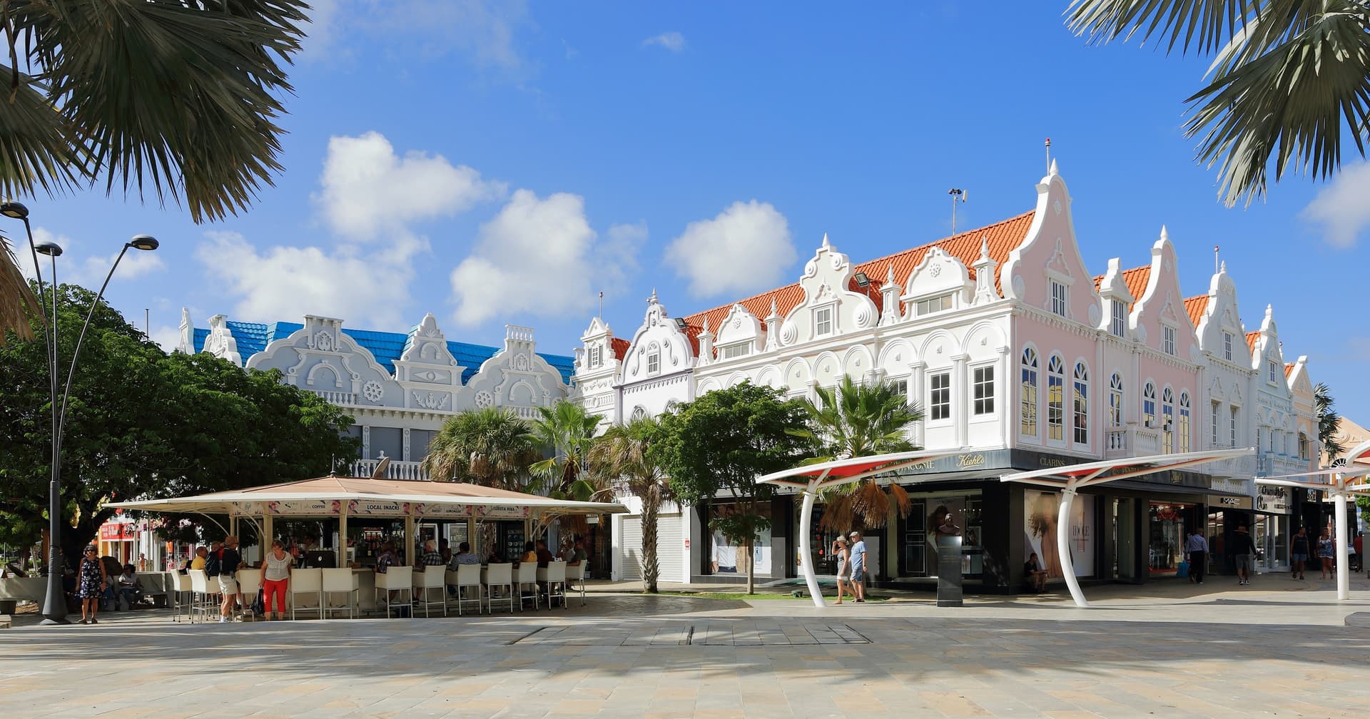 Witte en roze panden met sierlijke gevels in Nederlands-koloniale stijl aan het Plaza Daniel Leo in Oranjestad, Aruba, omlijst door palmbladeren