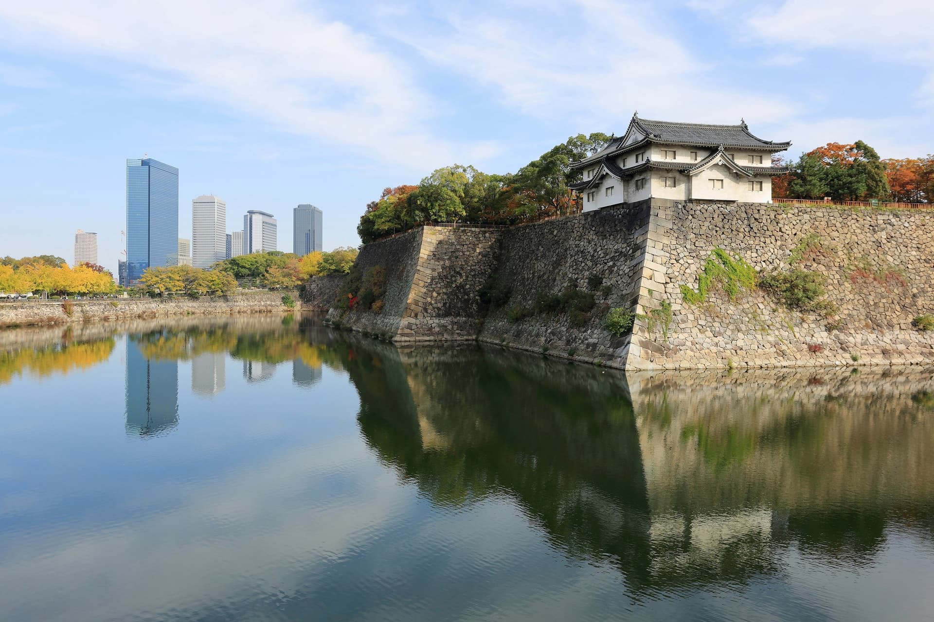 De hoge stenen muren en een wachttoren van kasteel Osaka weerspiegeld in de buitengracht, met herfstbomen en de torens van Osaka Business Park links