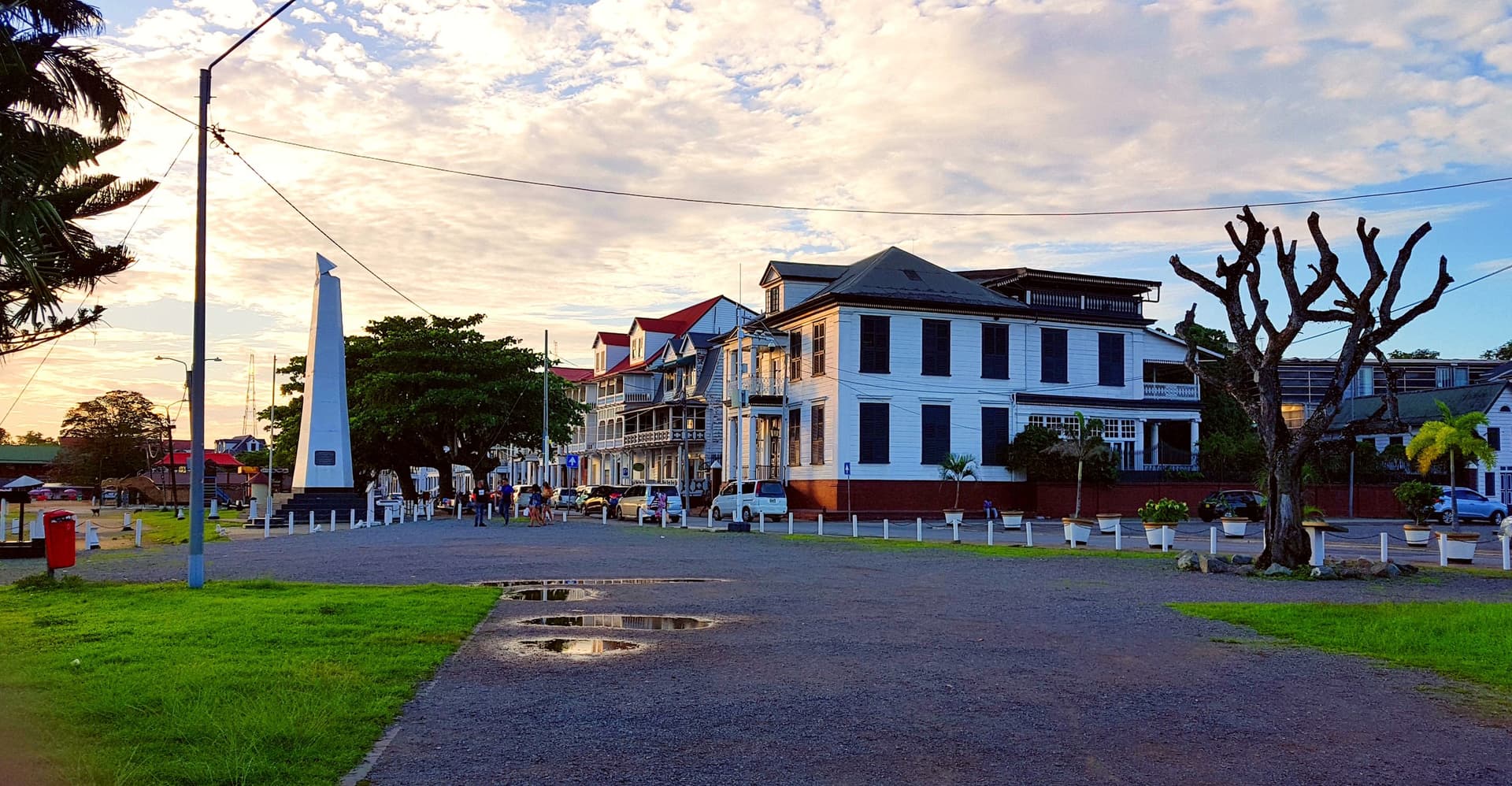 Witte houten koopmanshuizen met rode daken aan de Waterkant in Paramaribo bij een zonnige namiddag, met een obelisk en palmbomen