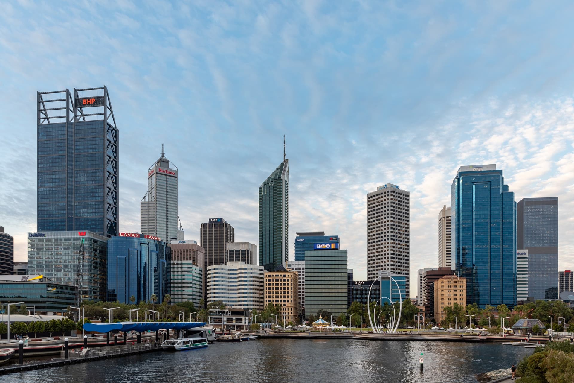 De skyline van Perth boven de jachthaven van Elizabeth Quay, met de witte Spanda-sculptuur aan het water en schapenwolkjes in de lucht