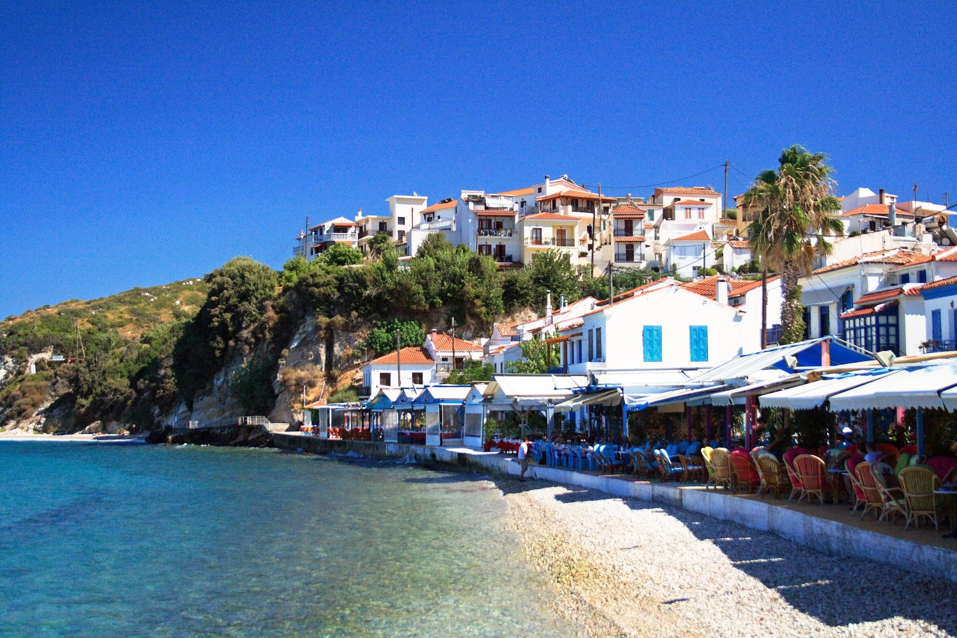 De haven van Kokkari op Samos met terrasjes aan het kiezelstrand, witte huizen met rode daken en palmen onder een strakblauwe lucht