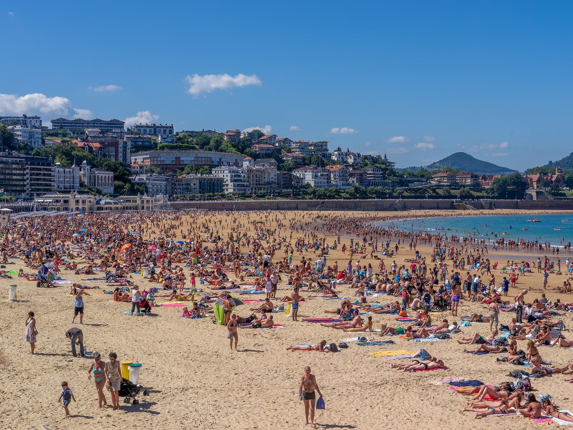 Het drukke stadsstrand van La Concha in San Sebastián op een zonnige zomerdag, met de stad en groene heuvels erachter