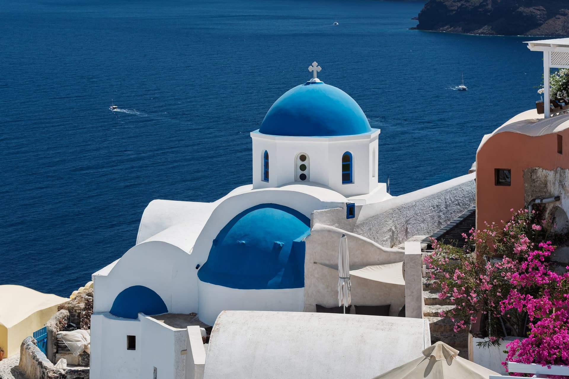 Een witte kerk met blauwe koepels boven de caldera van Santorini, met roze bloemen en de diepblauwe zee op de achtergrond