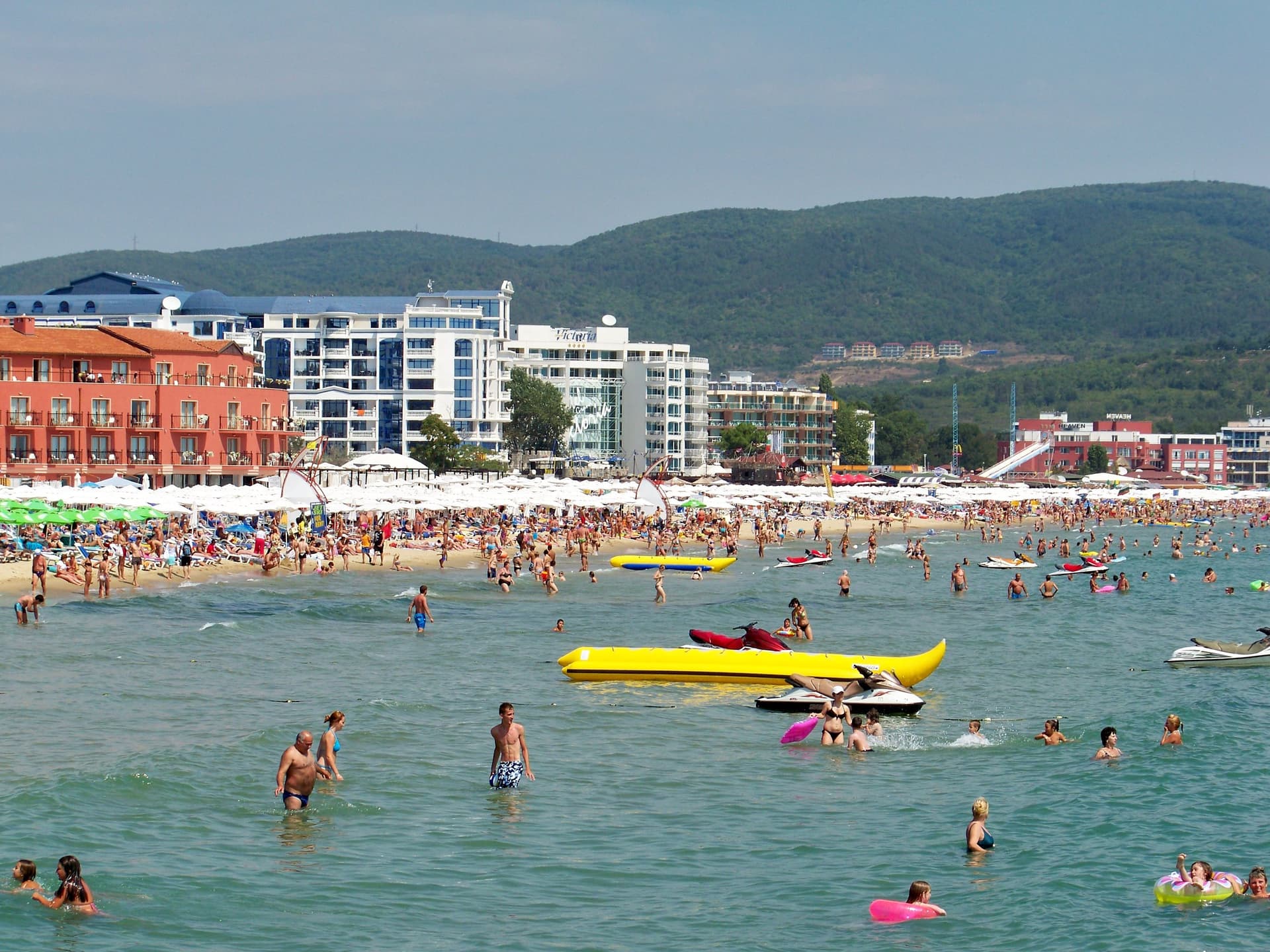 Druk zandstrand van Sunny Beach met badgasten in zee, gele bananenboten en hotels onder de groene heuvels van het Balkangebergte