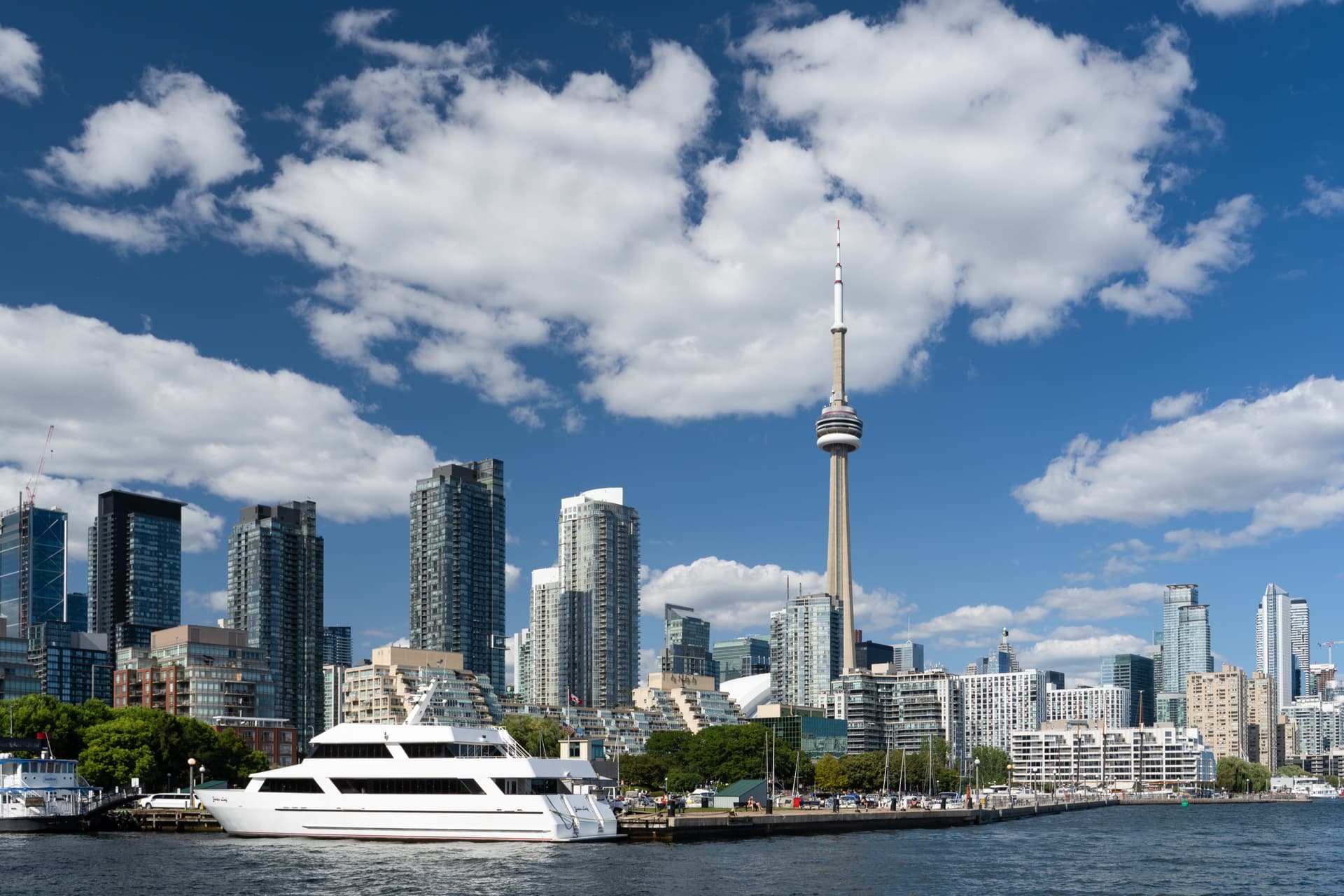 De CN Tower en woontorens aan de waterkant van Toronto onder een blauwe lucht met stapelwolken, gezien vanaf Ireland Park met een rondvaartjacht op het Ontariomeer