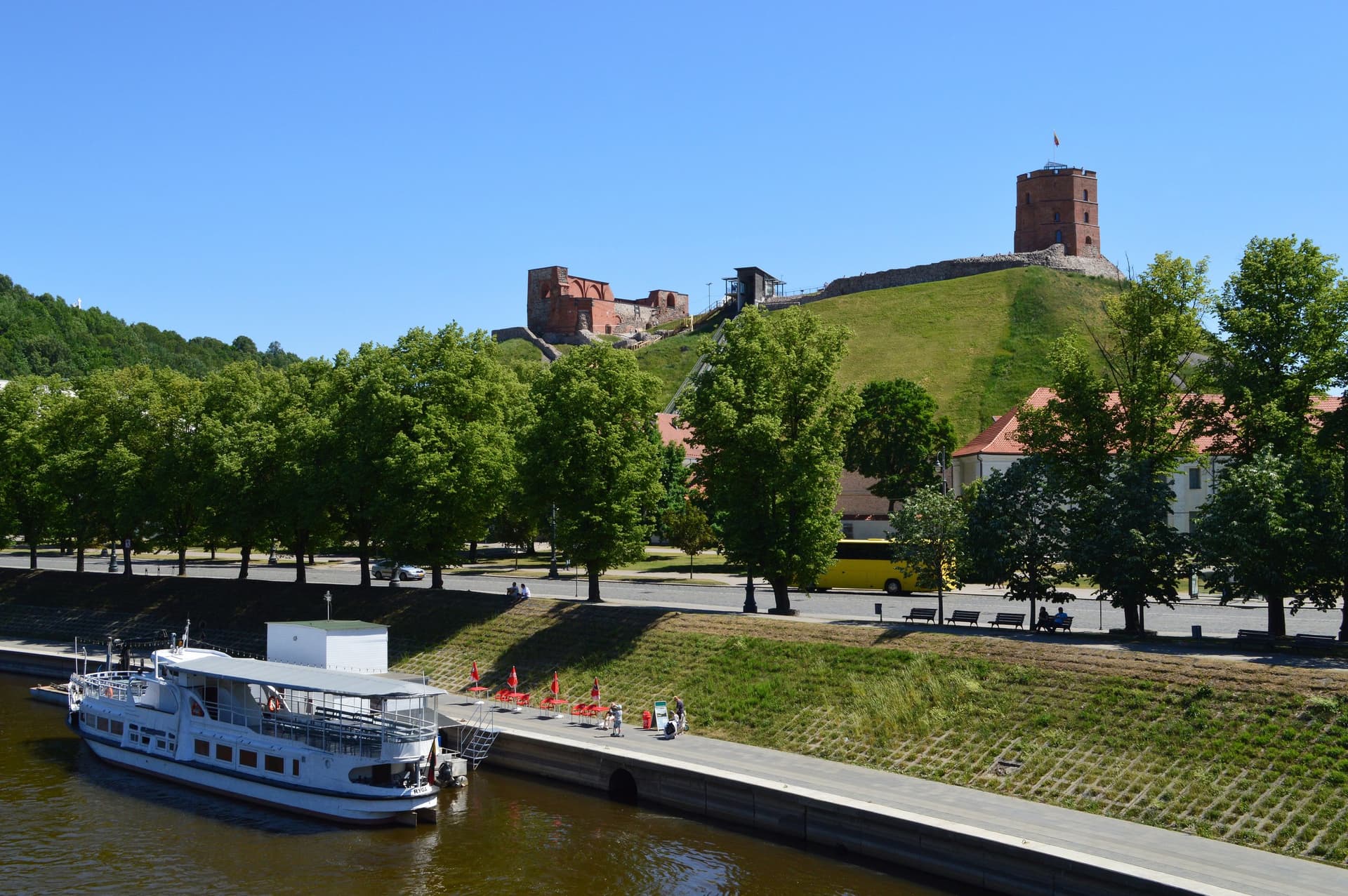 De Gediminastoren op de groene burchtheuvel boven de rivier in Vilnius, met een rondvaartboot aan de kade